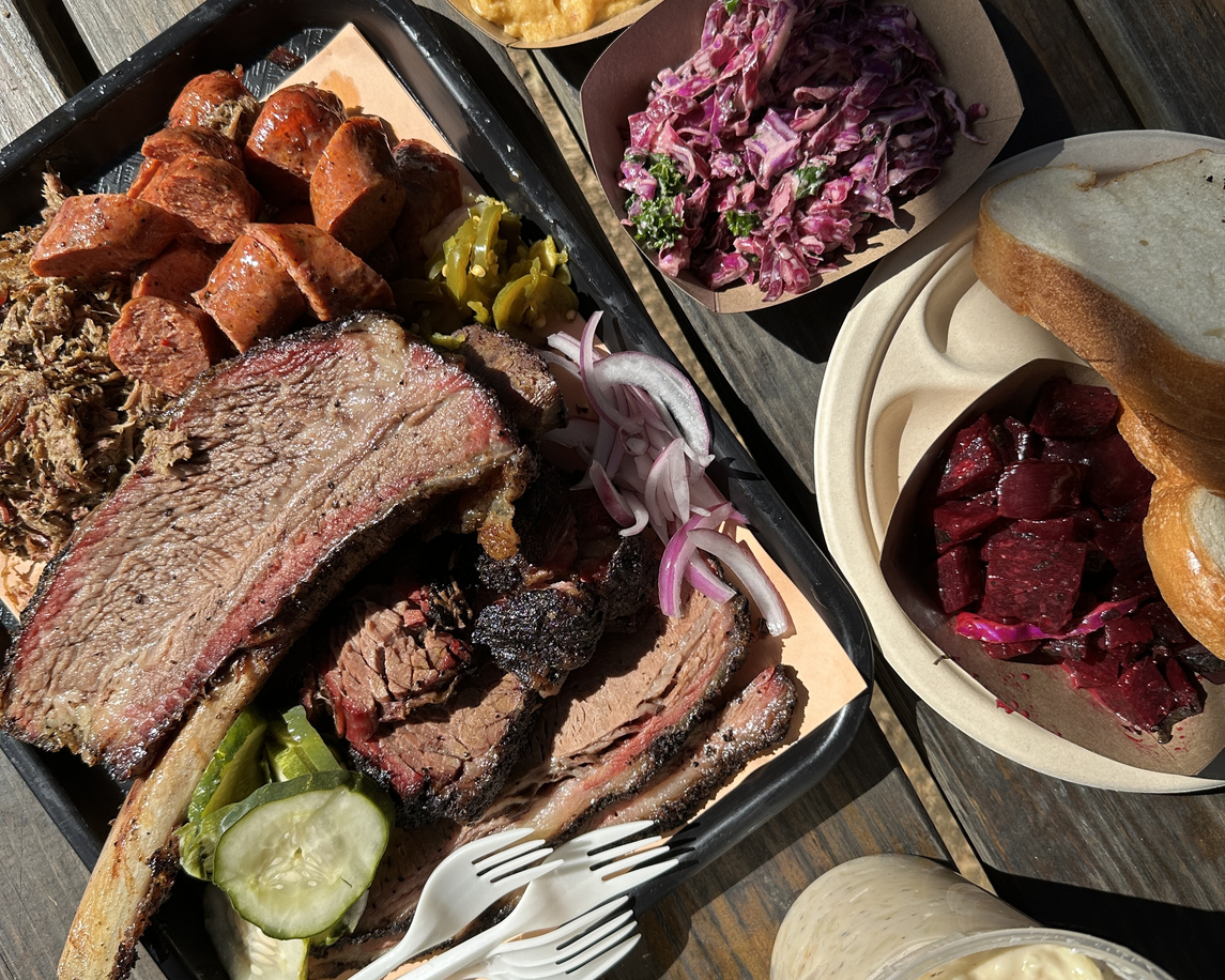An overhead view of a large tray of Texas barbecue foods, including beef rib, beef brisket, sausage, pulled lamb, plus sides of pickles, mac and cheese, grits, beets, bread, and more