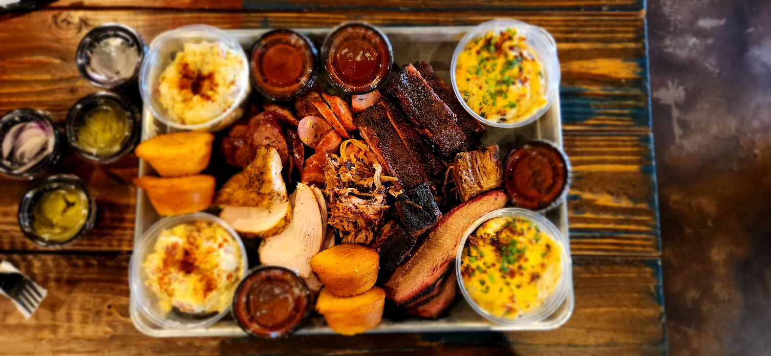 Large silver tray layered with variety of BBQ meats, sides, rolls, and sauces on a wood table