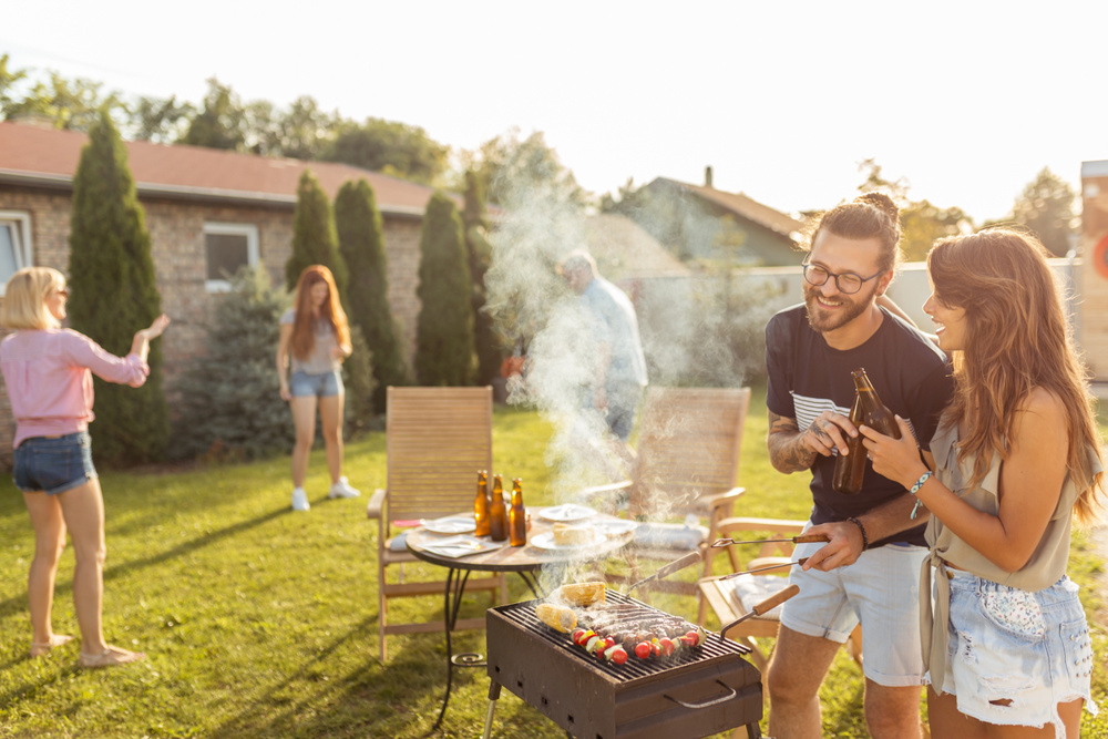 Group of cheerful young friends having a backyard barbecue party, grilling meat, making a toast, drinking beer and having fun on a sunny summer day