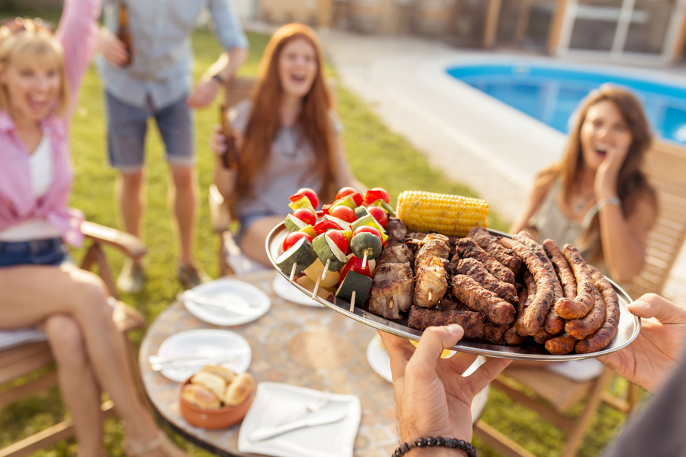 Group of cheerful young friends gathered around the table, drinking beer and having fun at a backyard poolside barbecue party, host bringing plate of grilled meat and vegetables