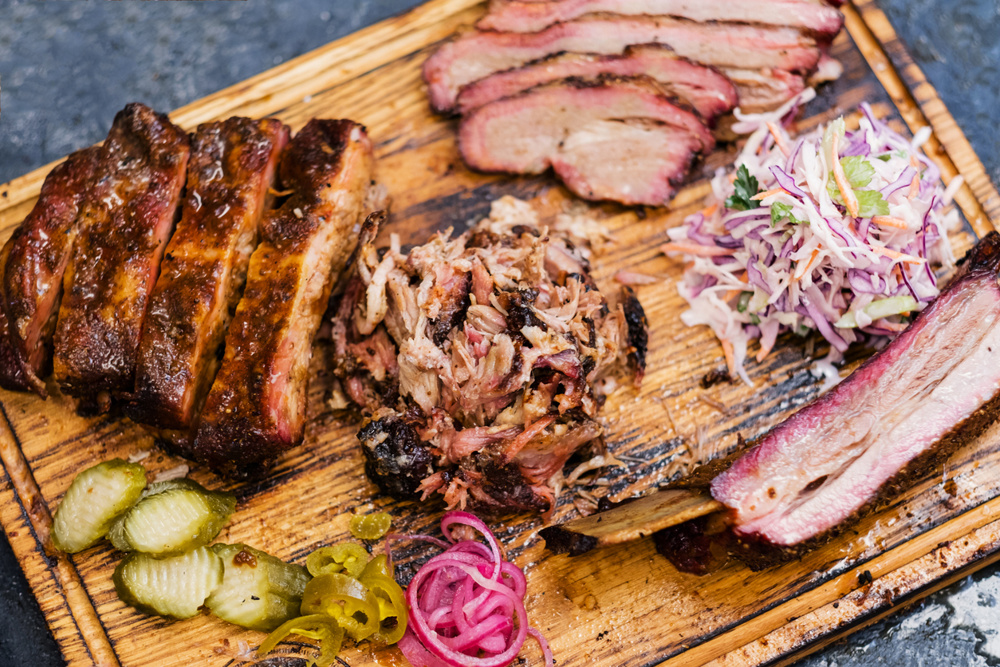 Smoked meat assortment on wooden board. Top view of sliced beef brisket, pulled pork, ribs, coleslaw salad, pickles