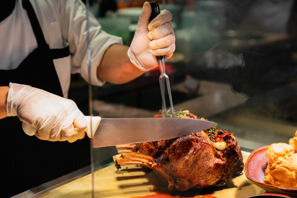 A chef wearing gloves and a black apron slices a juicy prime rib roast using a carving fork and a large knife. The scene takes place at a buffet or carving station, showcasing professional food preparation and hospitality service. Rich textures and warm lighting highlight the perfectly roasted meat